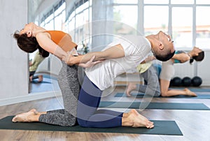 Lovers of active lifestyle perform pair yoga supported backbend on knee in gym
