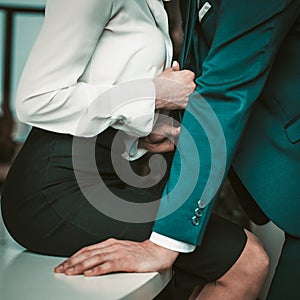 Lovers couple at workplace. Woman in formalwear sits on a table and pulls a man for his tie. Passionate affair in the