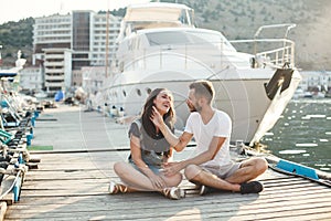 Lovers, guy and girl, are sitting on a wooden pier, holding hands and laughing