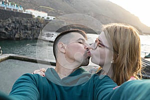 Lovers kissing with passion taking selfie in natural pool. Caucasian blonde girlfriend with glasses kiss latin boyfriend at sunset