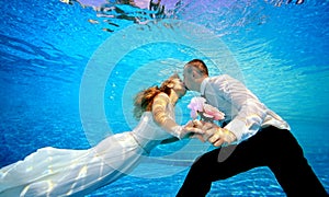 Lovers man and woman in wedding dresses kissing underwater in the pool and holding flowers in his hand