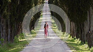 Lovers walk along cypress alley, guy hugs, kisses girlfriend in summer red dress