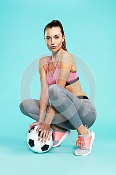 She loves football. Full length of young and sexy woman in sportswear squatting with soccer ball against blue background