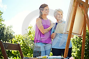 She loves watching her mother paint. a senior woman and her adult daughter painting in a park.