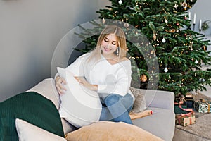 Lovey woman is sitting on a gray sofa near the Christmas tree in the living room and holding a soft pillow in her hands and