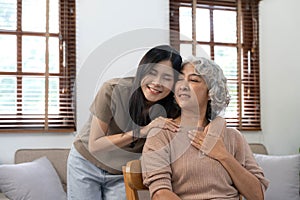 Loving adult daughter hugging older mother, standing behind couch at home, family enjoying tender moment together, young