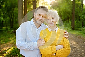 Loving adult son tenderly embracing elderly mother during walking at summer park. Two generations of family. Meeting of mom and