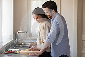 Loving affectionate millennial newlyweds preparing meal together at home.