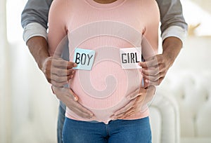 Loving African American couple holding BOY and GIRL cards near pregnant belly, close up view