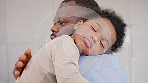 Loving African American dad embracing baby and baby girl sleeping on stepfather shoulder. Happy family with pleasure spending time