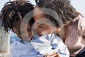 Loving african american mother kissing her little son watching video or playing game using mobile phone outdoors. Caring mother