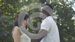 Loving African American young man adjusting hair of smiling shy Caucasian woman and hugging lover outdoors. Side view