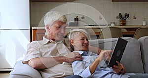 Loving aged couple cuddle on couch use modern digital pad