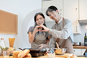 A loving Asian couple is making a heart hand sign together while making pancakes in the kitchen