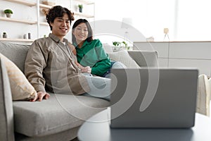 Loving Asian couple watching movie on laptop, sitting on couch at home, free space