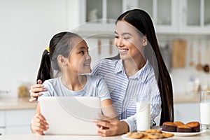 Loving asian mother and her daughter using digital tablet, looking for new recipes while sitting at kitchen table