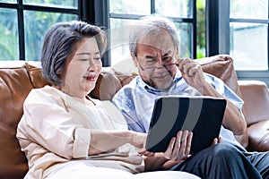 Loving Asian Senior elderly woman and old man couple sit on sofa in living room use laptop to read news and online video call with