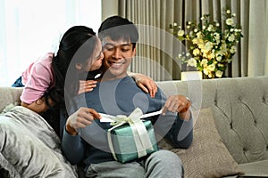 Loving Asian wife giving present box to her husband and kissing on cheek. Love, relationship and lifestyle concept