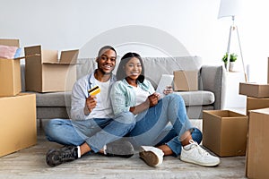 Loving black couple with digital tablet and credit card choosing furniture online after moving to new apartment