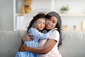 Loving black girl in pretty dress hugging her mother on sofa in living room. Strong family relationships concept