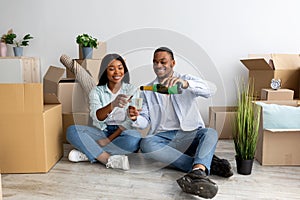 Loving black spouses celebrating moving day with champagne, sitting on floor in new apartment among cardboard boxes