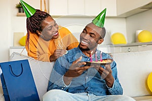 Loving black wife making surprise for her husband, happy man holding birthday pie while sitting on sofa at home