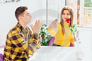 Loving boy and girl sitting in cafe, girl looking happy, taking selfie with flower, while boy doesn`t understand why she
