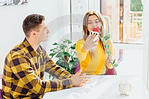 Loving boy and girl sitting in cafe, girl looking happy, taking selfie with flower, while boy doesn`t understand why she