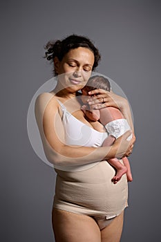 Loving caring multiethnic young woman mother carrying and hugging her newborn baby, isolated over gray studio background