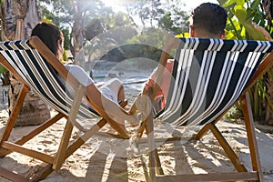 Loving caucasian young couple holding hands and relaxing on deck chairs at beach