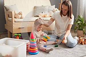 Loving Caucasian young mother and cute toddler girl playing with colorful blocks, sitting on warm floor at home, happy mum and