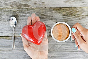 Loving coffee concept with hands of a woman holding one red heart and one cup of expresso coffee on wooden background