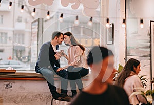 Loving couple dressed in sweaters and jeans is sitting close to each other on the windowsill in a cafe and holding cups