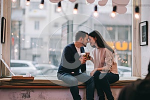 Loving couple dressed in sweaters and jeans is sitting close to each other on the windowsill in a cafe and holding cups