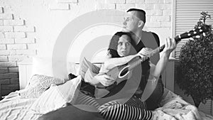 Loving couple with guitar sitting on bed. Man hugging girlfriend playing blue acoustic guitar while sitting on bed