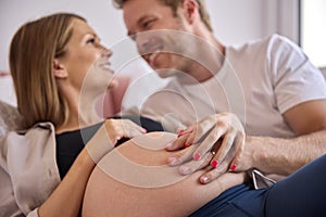 Loving Couple At Home Lying On Bed With Man Touching Pregnant Woman's Stomach 