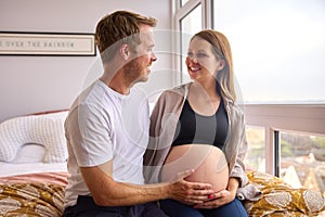 Loving Couple At Home Sitting On Bed With Man Touching Pregnant Woman's Stomach 