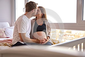 Loving Couple At Home Sitting On Bed With Man Touching Pregnant Woman's Stomach 