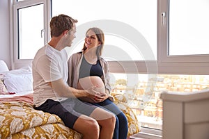 Loving Couple At Home Sitting On Bed With Man Touching Pregnant Woman's Stomach 