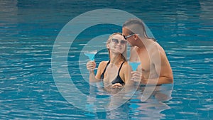 The loving couple hugs and kisses, drinking blue cocktail alcohol liquor in swimming pool at hotel outdoor. Portrait of