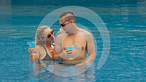 The loving couple hugs and kisses, drinking blue cocktail alcohol liquor in swimming pool at hotel outdoor. Portrait of