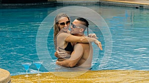 The loving couple hugs and kisses, drinking blue cocktail alcohol liquor in swimming pool at hotel outdoor. Portrait of