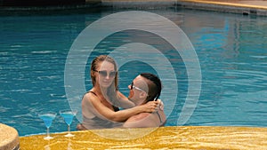 The loving couple hugs and kisses, drinking blue cocktail alcohol liquor in swimming pool at hotel outdoor. Portrait of