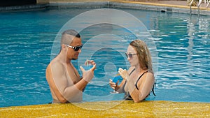 The loving couple hugs and kisses, drinking blue cocktail alcohol liquor in swimming pool at hotel outdoor. Portrait of