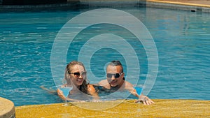 The loving couple hugs and kisses, drinking blue cocktail alcohol liquor in swimming pool at hotel outdoor. Portrait of