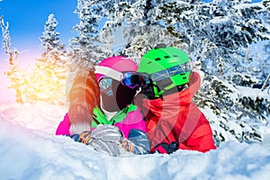 Loving couple kisses in winter snow, snowboarder and skier lie against background of forest, sunny day