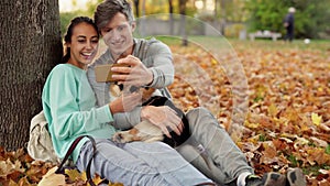 Loving Couple making selfie on smartphone with cute tricolor Welsh Corgi Dog In City Park hugging relaxing enjoying warm