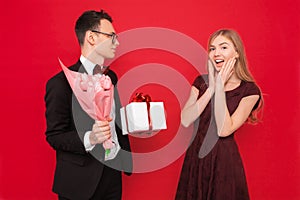 A loving couple, a man gives a shocked woman a bouquet of tulips and a box with a gift on a red background. Valentine`s Day