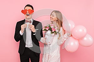 A loving couple, a man holding two paper hearts in his eyes, and a woman holding a bouquet of flowers, on a pink background