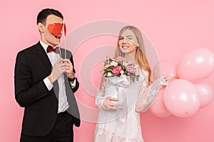 A loving couple, a man holding two paper hearts in his eyes, and a woman holding a bouquet of flowers, on a pink background